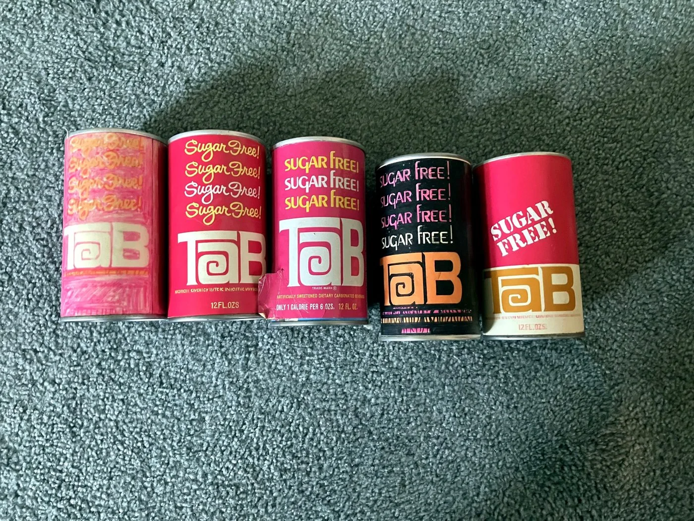 Five vintage TaB cans in a row showing the redesign's progression, from a hand-marker concept with sugar free repeated four times to the finished pink production can.